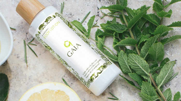 A bottle of Gaia body lotion, next to a sprig of mint, rosemary leaves and lemon.