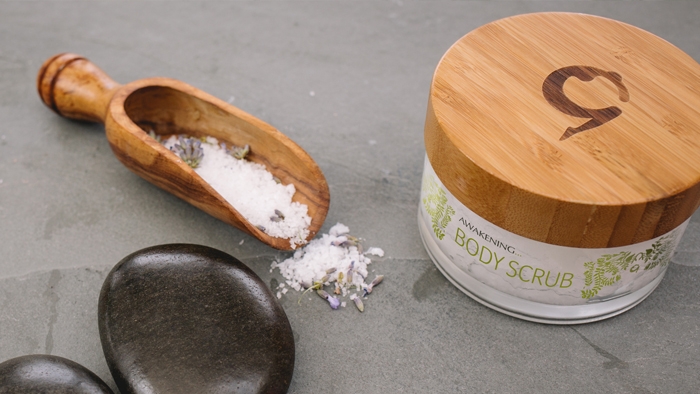 A pot of Gaia body scrub beside a wooden scoop of salt and lavender. 