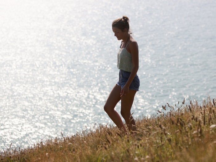 A woman walks down a hill with the sea behind her.