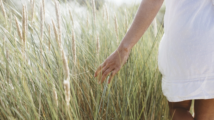 A woman brushes her hand against long grass as she walks through a field.